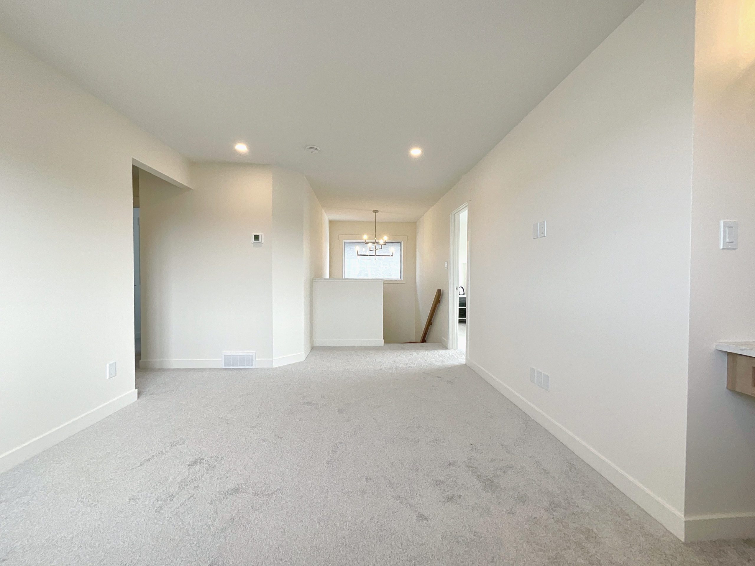 An empty bonus room with white walls and carpet flooring, shows the stairwell to main level and chandelier.