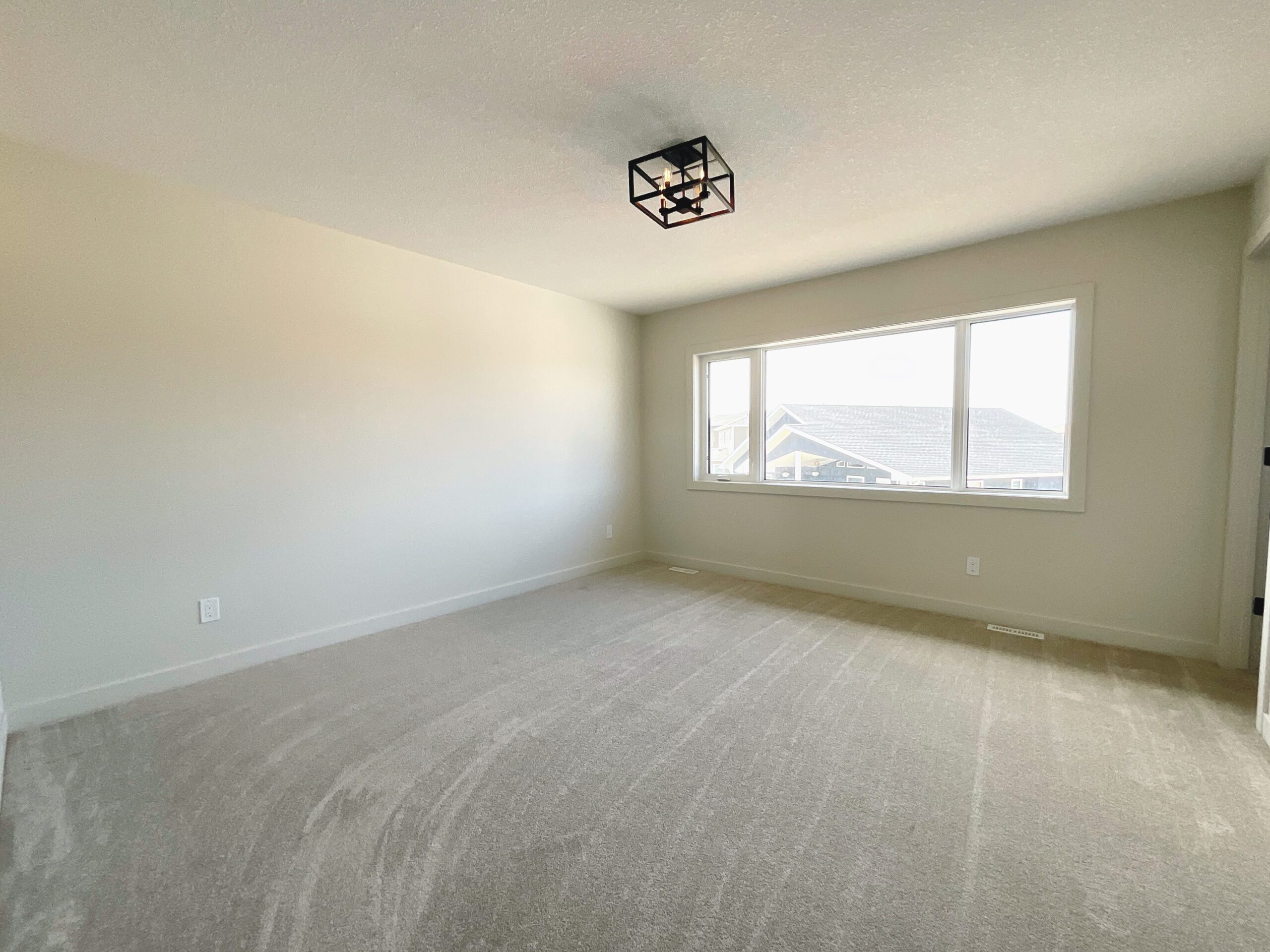 An empty primary bedroom with a large window and a semi-flush light fixture with carpet flooring.