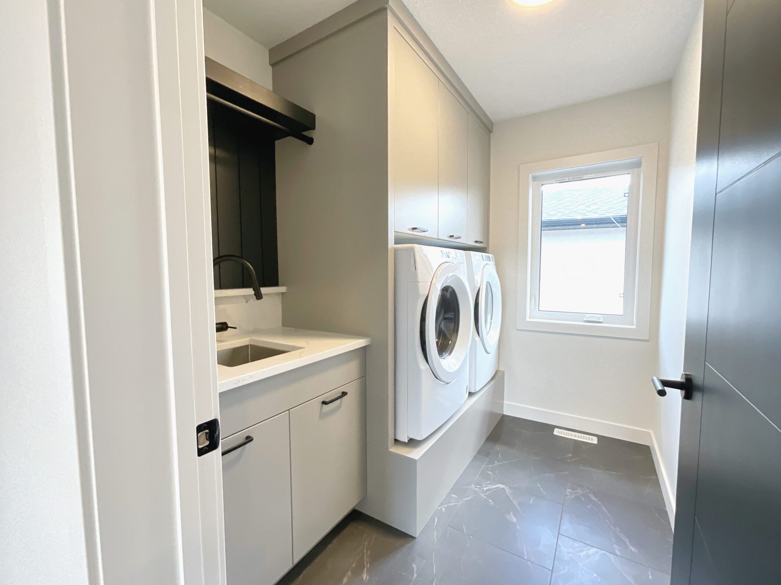 A laundry room with built-in pedestal cabinetry for the washer and dryer with a sink and closet rod and tiled flooring.