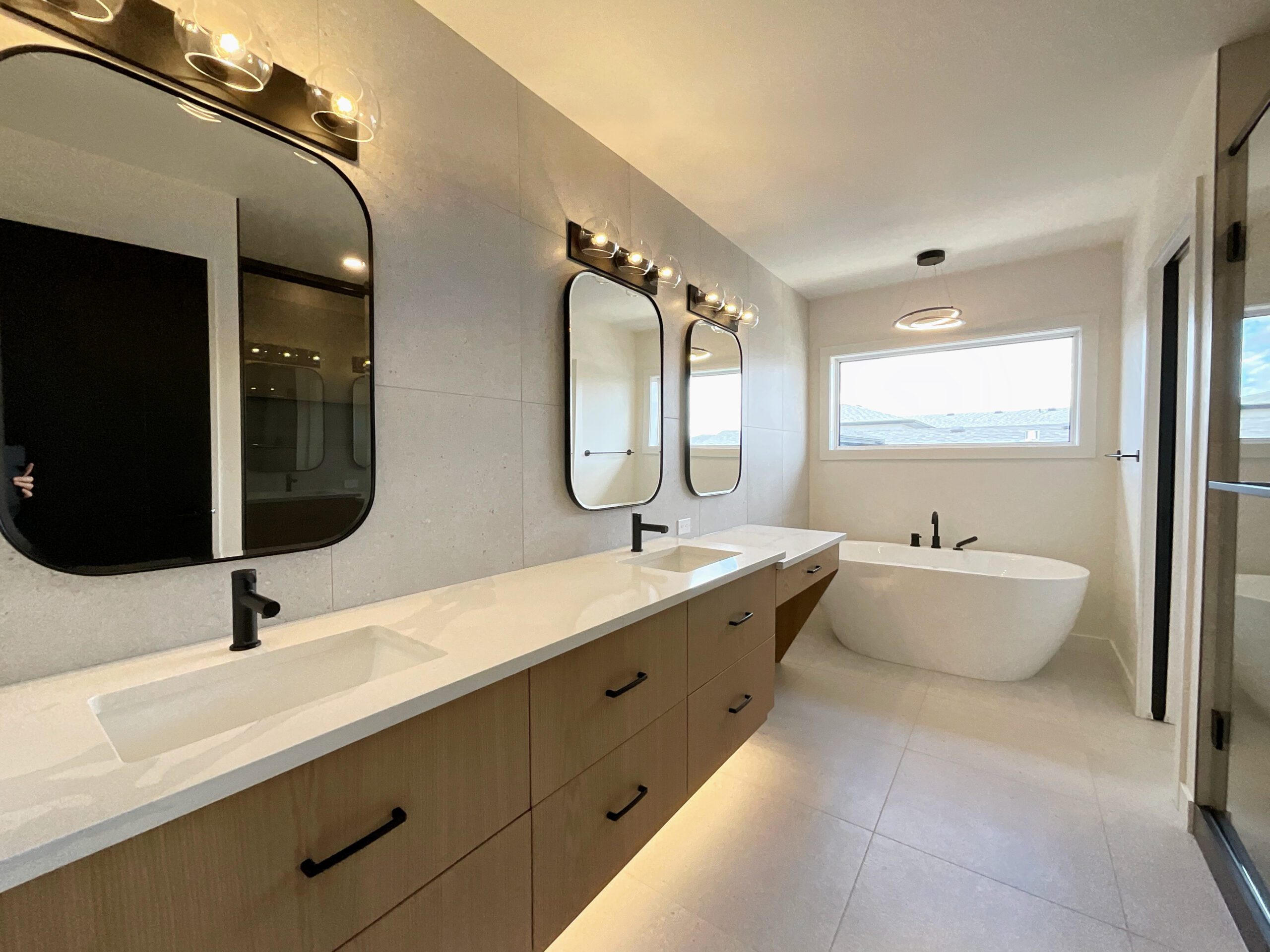 The primary ensuite bathroom with double sinks, makeup counter, oval mirrors, a freestanding tub with modern chandelier above, and tiled flooring which also serves as a backsplash running the whole wall of the vanity.