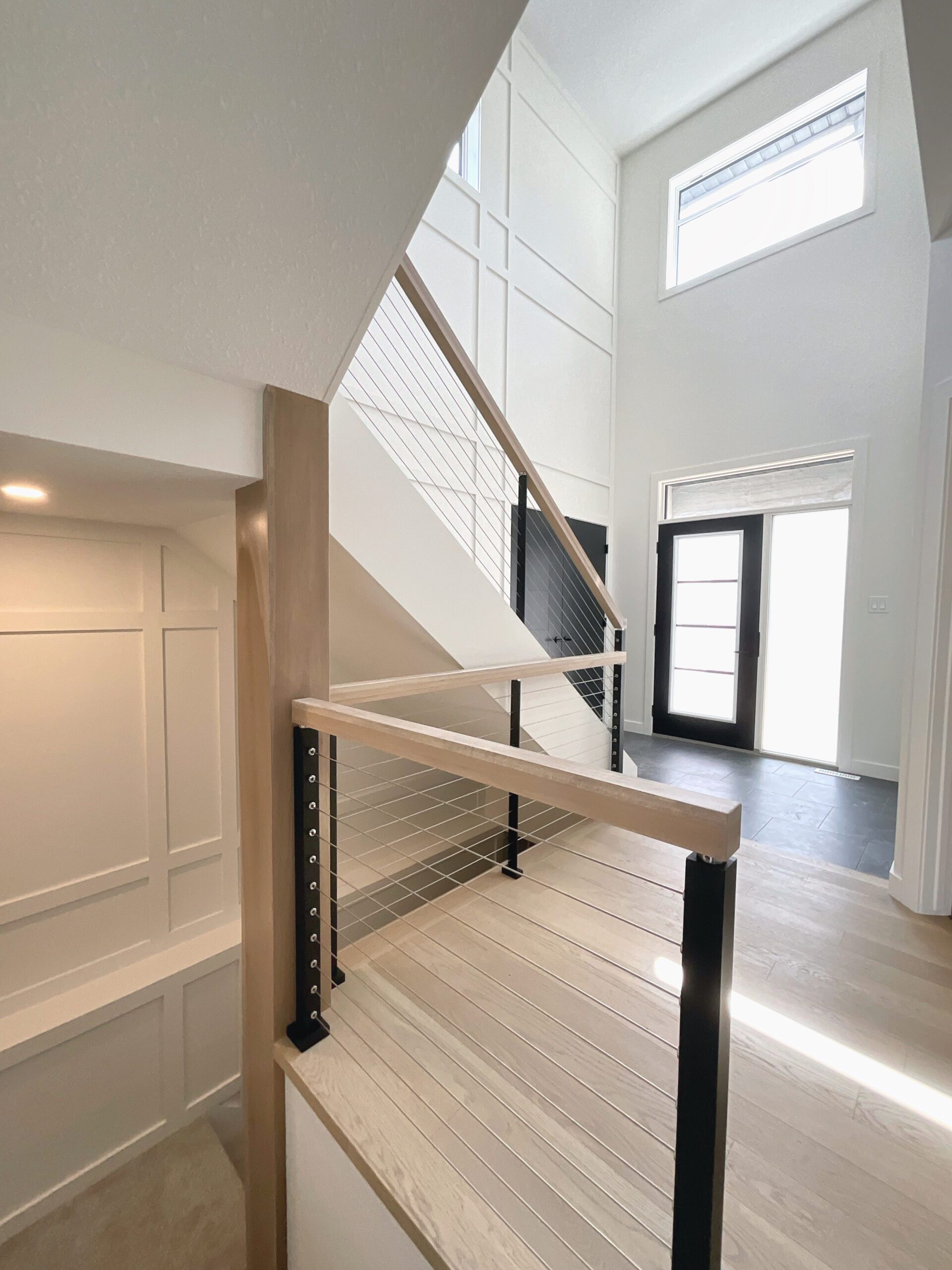 A staircase leading to second and basement level with decorative trim on the wall, cable railing and hardwood flooring, also shows front door entryway with tile flooring.