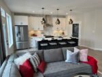 Modern two-storey home in Pilot Butte with white cabinetry and a central island overlooked by pendant lights, adjacent to a living area with a gray sofa adorned with colorful cushions.