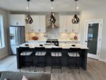 Modern kitchen interior in a two-storey home in Pilot Butte with a central island and bar stools, stainless steel appliances, and pendant lighting.