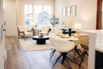 Modern living and dining area with wooden flooring, white chairs, a round dining table, and a coffee table. Large windows in the background bring in natural light.