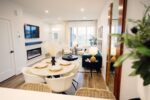 Modern living room with a dining area, fireplace, and wall-mounted TV. Features white walls, large windows, and a seating area with a green sofa and round coffee table.