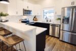 Modern kitchen with white cabinets, stainless steel appliances, a black sink, and wood flooring. Bar stools are placed at the kitchen island.