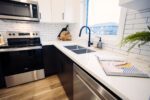 Modern kitchen with stainless steel appliances, white tile backsplash, dual sinks, and a white countertop. A plant, cutting board, and magazine are on the counter. Wooden floor visible.