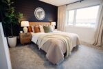 Modern bedroom with a bed, layered blankets, and pillows. A round mirror is on the dark accent wall. A potted plant and a window brighten the room. Two lamps sit on wooden nightstands.