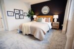 Stylish bedroom with a bed, neutral bedding, black accent wall, framed artwork, lamps, and a round mirror. A plant is in the corner, and a window is on the right with curtains.