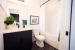 A modern bathroom with a dark vanity, white countertop, toilet, bathtub with a shower curtain, potted plant, and artwork on the wall.