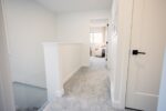 A carpeted hallway with white walls and doors leading to a bright room with a window at the end.