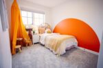 A cozy bedroom with a single bed, beige and white bedding, an orange wall accent, and a yellow canopy by the window. A small side table and chair are near the bed.