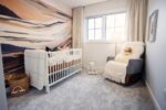 A cozy nursery with a crib, a rocking chair with a teddy bear, and a basket. A landscape mural covers one wall, and a lamp is lit by the window with white curtains.