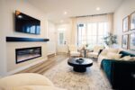 A modern living room with a wall-mounted TV, electric fireplace, green sofa, round black coffee table, and four white chairs. Large windows and hardwood floors brighten the space.