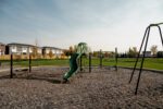 A playground with a green slide, swings, and play structures on a gravel surface, surrounded by grassy areas and nearby houses.