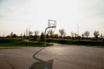 Outdoor basketball court with a single hoop, surrounded by trees and a paved path. The sun casts long shadows across the court.