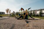 A playground with slides, climbing structures, and swings on a mulch surface, surrounded by grass, trees, and nearby residential buildings under a clear sky.